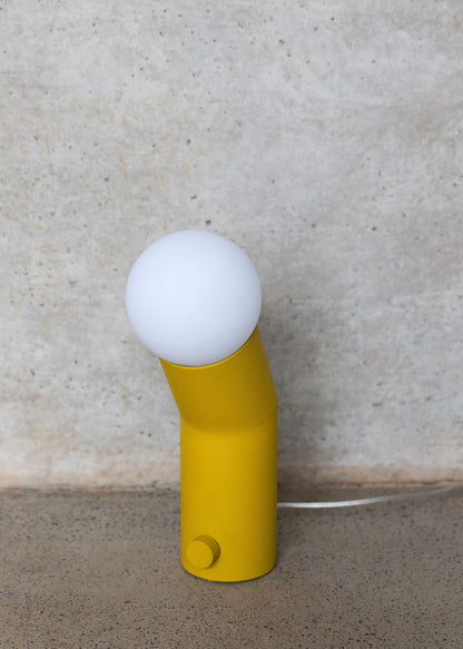 A yellow capsule-shaped lamp with a white glass shade, positioned on a concrete surface against a wall, with a clear cable and a two-pin plug.